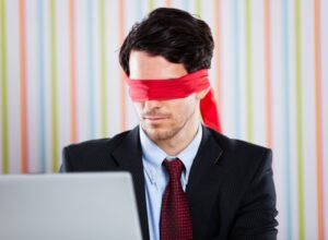 Businessperson in suit and tie wearing a red blindfold while working on a laptop, symbolizing financial decision-making without visibility