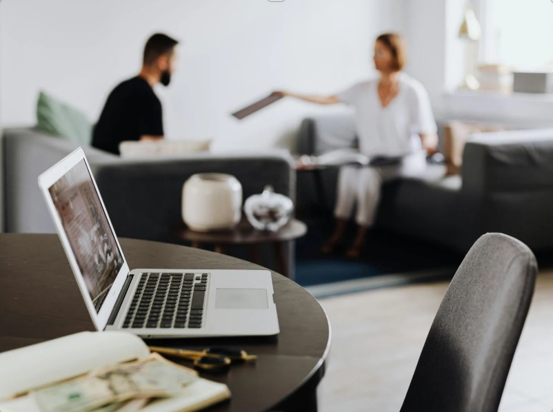 Two people seated in a modern office setting during a client onboarding meeting, with a laptop, notebook, and currency notes on the table in the foreground.
