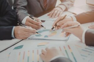 Group of business professionals gathered around a table reviewing pie charts, bar graphs, and financial documents during a collaborative planning meeting