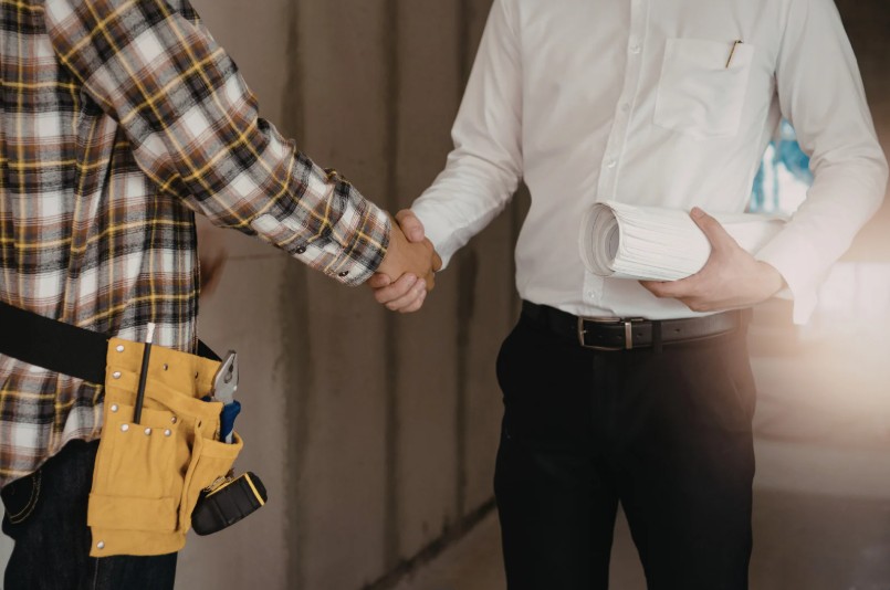 A contractor in a tool belt shakes hands with a professional holding building plans inside an unfinished construction space. Description