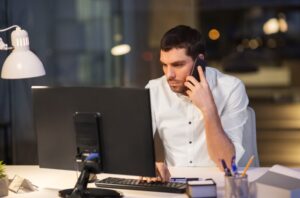 Business owner sitting at a desk at night, reviewing financial information on a computer while speaking on the phone.