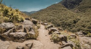 A dirt mountain trail winding between large rocks and green vegetation, gradually leveling out under a clear sky.