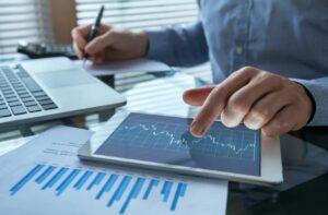 A person pointing at financial charts on a tablet screen while taking notes, with printed reports and a laptop visible on the desk