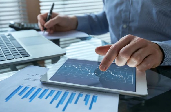 A person pointing at financial charts on a tablet screen while taking notes, with printed reports and a laptop visible on the desk