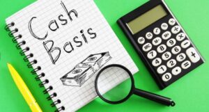 Notebook reading "Cash Basis" with a hand-drawn stack of cash, next to a calculator and magnifying glass on a green background