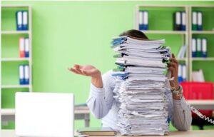 A person sitting at a desk with one hand raised in a helpless gesture, hidden behind an enormous stack of papers and files, with green shelving and binders in the background.
