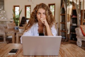 A woman with curly hair sitting at a wooden table, hand raised to her mouth in a worried expression, staring intently at an open laptop
