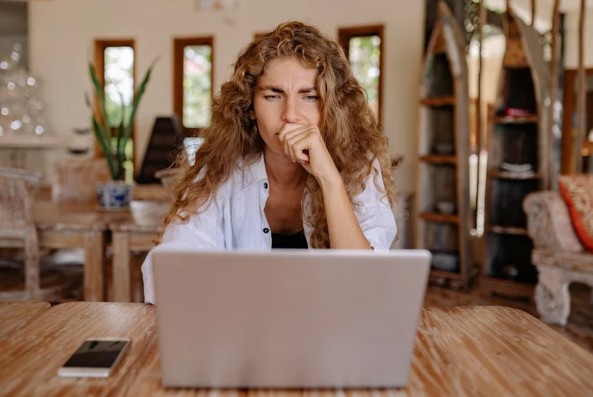 A woman with curly hair sitting at a wooden table, hand raised to her mouth in a worried expression, staring intently at an open laptop