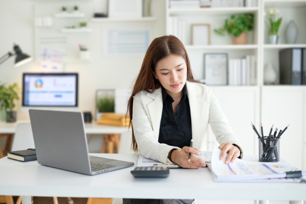 A woman in a white blazer sitting at a clean office desk, reviewing printed financial charts alongside an open laptop and calculator.
