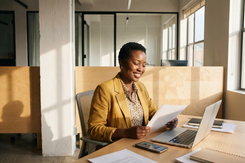 A woman in a gold blazer sitting at a wooden desk, smiling as she reviews a printed document, with a laptop and phone visible in a bright, naturally lit office.