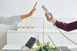 A hand in a polka dot sleeve gesturing stop toward a corded phone handset being offered across a desk, with filing cabinets and laptops in the background.