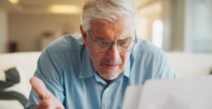 An older man with glasses leaning forward with a puzzled expression, hand raised in a questioning gesture, reviewing printed documents in a warmly lit room.