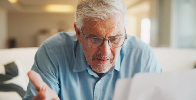 An older man with glasses leaning forward with a puzzled expression, hand raised in a questioning gesture, reviewing printed documents in a warmly lit room.
