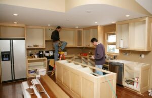 Two contractors installing wood cabinets in a kitchen renovation, with one worker on a ladder and the other reviewing plans near the island frame.