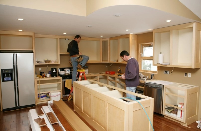 Two contractors installing wood cabinets in a kitchen renovation, with one worker on a ladder and the other reviewing plans near the island frame.
