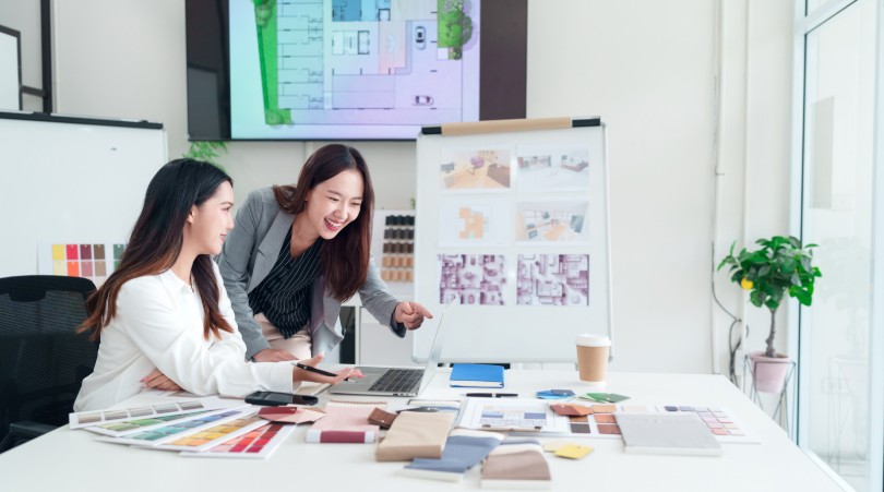 Two interior designers smiling and collaborating at a table covered with fabric swatches and material samples, with floor plans displayed on a screen and design boards in the background.