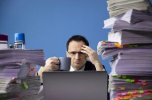 A man in glasses holding a coffee cup with a stressed expression, seated between two enormous stacks of tabbed and flagged paperwork in front of a laptop.