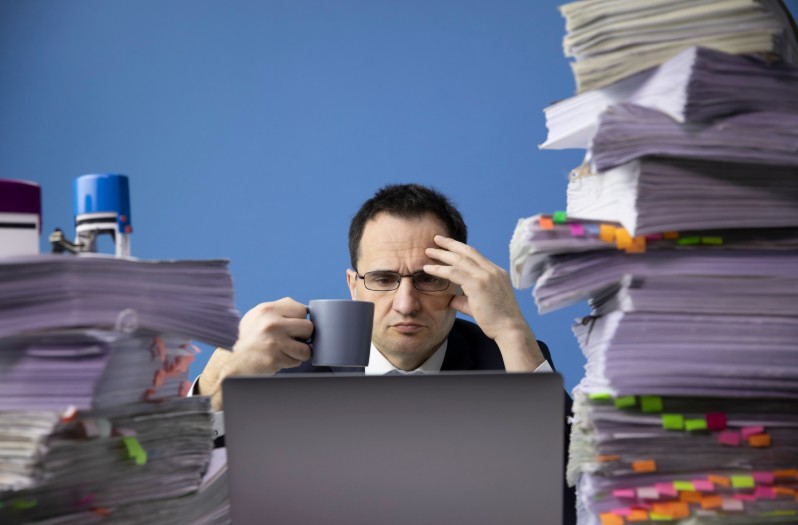 A man in glasses holding a coffee cup with a stressed expression, seated between two enormous stacks of tabbed and flagged paperwork in front of a laptop.