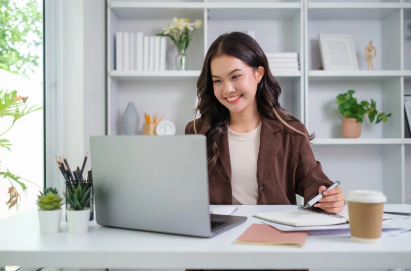 A young woman with long dark hair smiling at her laptop screen while taking notes, seated at a clean white desk in a bright, well-organized home office with shelving and plants in the background.